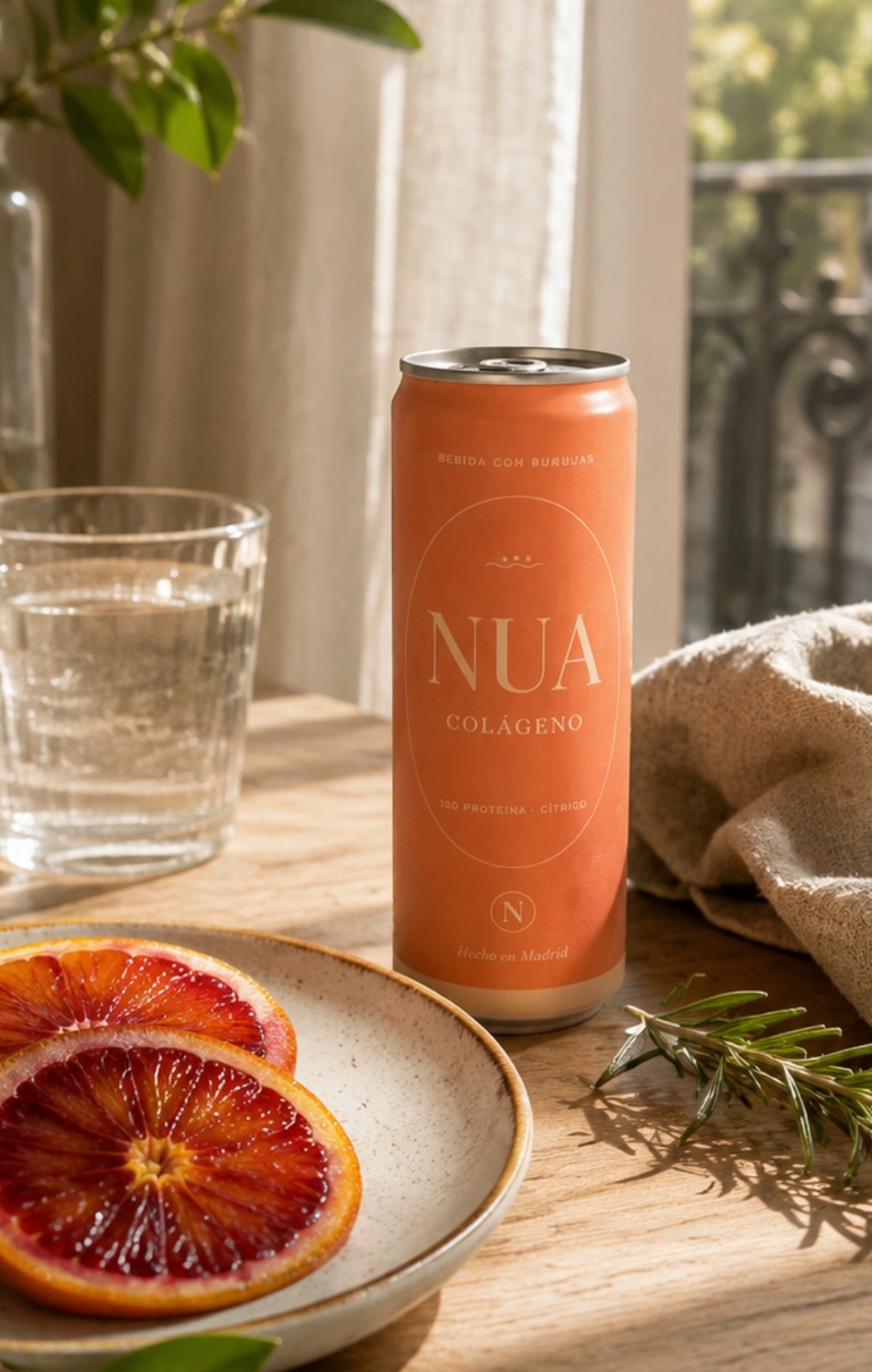 Una lata de NUA Colágeno sobre una mesa de madera, con un plato de naranjas sanguinas, un vaso de agua y romero.